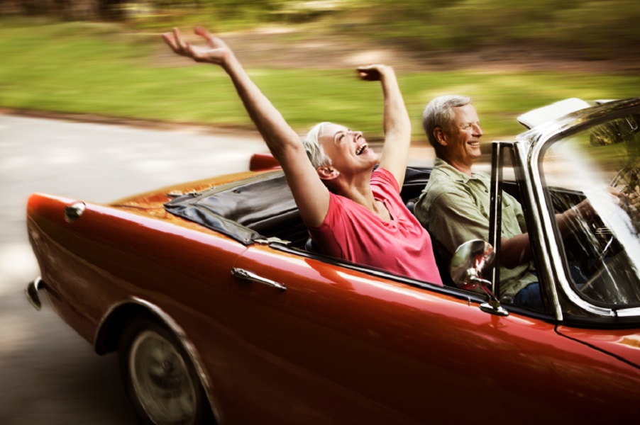 Senior woman throws her arms in the air while her husband drives her down a road in their convertible. Horizontal shot.